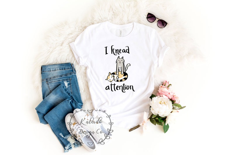May include: White t-shirt with a graphic of two cats and the text "I knead attention". The shirt is laid flat on a white fluffy surface with a pair of sunglasses, a pair of jeans, and a bouquet of pink and white flowers.