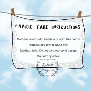 May include: A white fabric care label with black text hanging on a clothesline against a blue sky background. The label reads "FABRIC CARE INSTRUCTIONS" and includes instructions for washing, drying, and ironing the fabric. The label also includes the text "Kaleido Design Co."