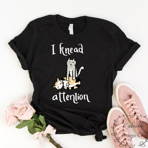 May include: Black t-shirt with a white graphic of two cats. The text on the shirt reads "I knead attention".