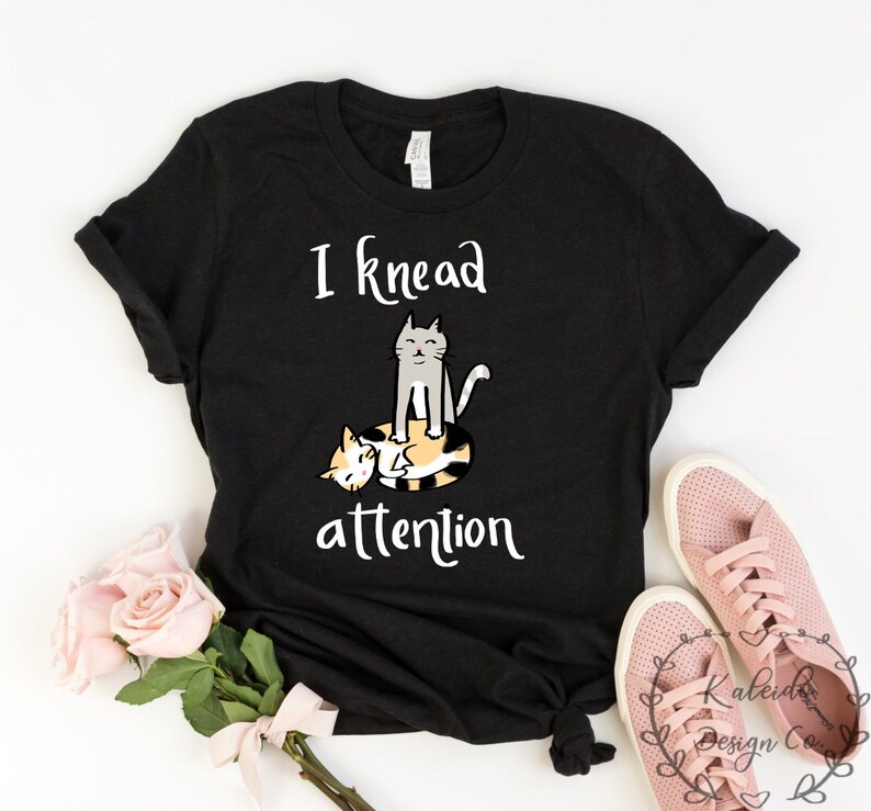 May include: Black t-shirt with a white graphic of two cats. The text on the shirt reads "I knead attention".