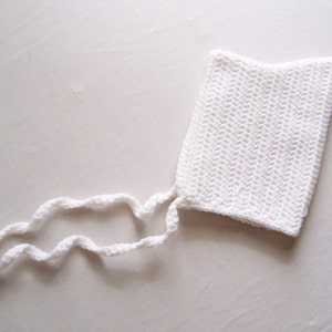 May include: A handmade, white crochet bonnet with two long, wavy ties. The bonnet is rectangular with a slightly curved top edge. The crochet stitch creates a textured surface. The ties are also crocheted and appear soft.