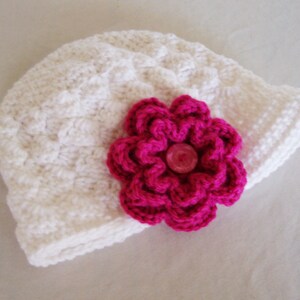 May include: A white crocheted hat featuring a textured, raised stitch pattern throughout the body. A large, layered fuchsia pink crocheted flower is attached to the side, centered with a small, round, translucent pink button detail.