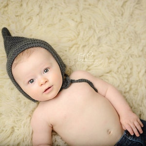 May include: A baby wearing a gray crocheted pixie bonnet and blue jeans, lying on a cream-colored, fluffy rug. The bonnet has a pointed top and ties under the chin. The baby is looking at the camera.