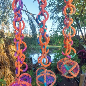 May include: Three colourful macrame art pieces hanging from a tree. Each piece is made of orange, purple, and blue rope, with a chain-like design of connected circles. The background shows a natural setting with a pond.