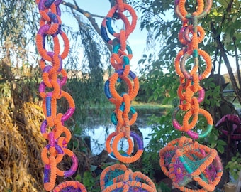 May include: Three hanging macrame art pieces in vibrant colors. Each piece features orange, purple, and blue rope, with a chain-like design of connected circles. The pieces are suspended from a tree branch, with a natural background.
