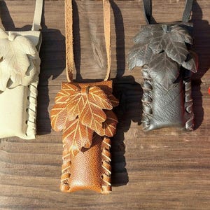 Könnte beinhalten: Fünf Lederbeutel mit Blattmotiven in verschiedenen Farben: Braun, Creme, Beige, Grau und Rot. Jeder Beutel hat einen Lederriemen und dekorative Nähte an den Seiten. Die Beutel werden auf einer Holzoberfläche präsentiert.