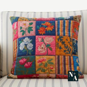 Puede incluir: Cojín cuadrado de patchwork con bordados florales. El cojín presenta una variedad de cuadrados coloridos, cada uno con un diseño floral diferente en tonos rosa, azul, naranja y amarillo. Sobre un fondo a rayas.