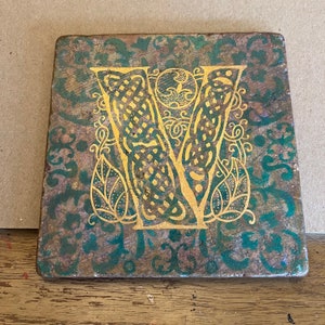 May include: A square, green and gold tile with a decorative letter V in the center. The letter is gold and has a Celtic knot design. The tile has a distressed, aged look.