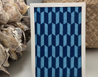 May include: A framed artwork with a geometric pattern in shades of blue. The design features a repeating chevron pattern in light and dark blue, set within a white frame. The artwork is displayed on a white surface.