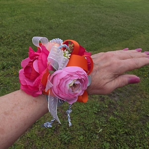 May include: A wrist corsage featuring pink and orange artificial flowers, accented with white ribbons and greenery. The floral arrangement is secured with a silver-colored wristlet, perfect for a special occasion.