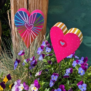 Könnte beinhalten: Zwei herzförmige Holzdekorationen, die in Pink, Rot und Weiß mit einem gestreiften und gepunkteten Muster bemalt sind. Die Herzen stehen in einem Blumenbeet mit lila und rosa Blüten.