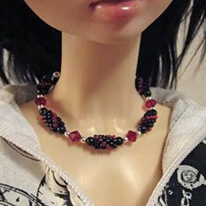 May include: A close-up of a doll wearing a black and red beaded choker necklace. The doll has long black hair and is wearing a white and black patterned shirt.