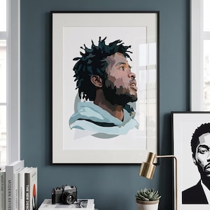 May include: A framed art print featuring a stylized portrait of a person with dark hair and a beard, wearing a light blue hoodie. The artwork is displayed on a dark blue wall, alongside other framed art and decorative items.