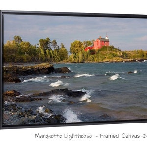 May include: A framed canvas print of a red lighthouse on a rocky shore with waves crashing against the rocks. The lighthouse is on a small island with trees and green grass. The sky is cloudy and the water is a dark blue. The print is 24x36 inches.