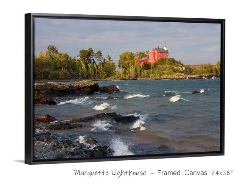 May include: A framed canvas print of a red lighthouse on a rocky shore with waves crashing against the rocks. The lighthouse is on a small island with trees and green grass. The sky is cloudy and the water is a dark blue. The print is 24x36 inches.