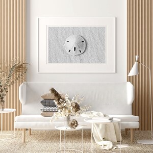 May include: A white couch with a white throw blanket and a white side table with a book on it. There is a vase with dried flowers on a white side table. A framed black and white photograph of a sand dollar on a sandy beach hangs on the wall above the couch.