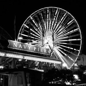 Op de afbeelding: Een zwart-witfoto van een reuzenrad op Navy Pier in Chicago. Het reuzenrad is verlicht en draait, en het bord voor Navy Pier is zichtbaar op de voorgrond.