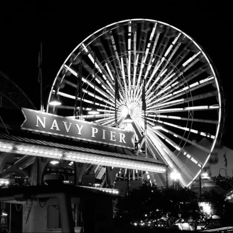 Op de afbeelding: Een zwart-witfoto van een reuzenrad op Navy Pier in Chicago. Het reuzenrad is verlicht en draait, en het bord voor Navy Pier is zichtbaar op de voorgrond.