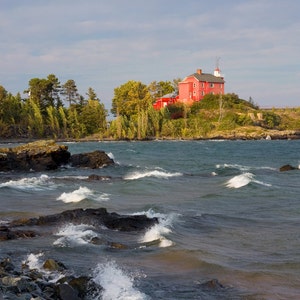 May include: A red lighthouse sits on a rocky outcropping overlooking a choppy lake. The lighthouse is a two-story building with a white roof and a small tower. The lake is a deep blue color and the waves are crashing against the rocks.