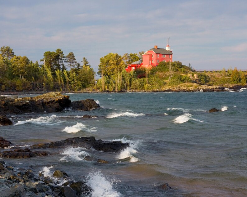 May include: A red lighthouse sits on a rocky outcropping overlooking a choppy lake. The lighthouse is a two-story building with a white roof and a small tower. The lake is a deep blue color and the waves are crashing against the rocks.