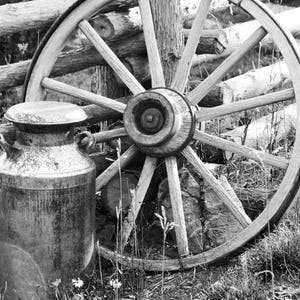 Puede incluir: Una foto en blanco y negro de una rueda de carro de madera vintage apoyada en una valla de madera. Una lata de leche de metal se encuentra delante de la rueda.