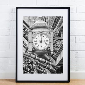 May include: Black and white photograph of a clock face with Roman numerals on a building facade. The clock is surrounded by intricate architectural details.
