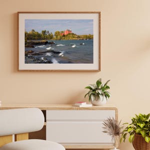 May include: A framed photograph of a red building on a rocky shoreline with blue water and white waves. The building is surrounded by trees and is located on a small island.