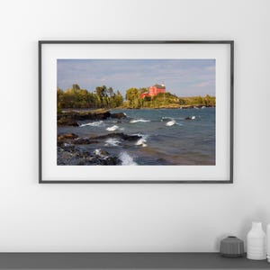 May include: A framed photograph of a red lighthouse on a rocky shoreline with blue water and white waves. The lighthouse is on a small island with trees in the background.
