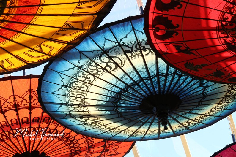 May include: A collection of colorful, patterned umbrellas hanging from the ceiling. The umbrellas are made of fabric and have intricate designs. The umbrellas are a variety of colors, including red, blue, yellow, and white.