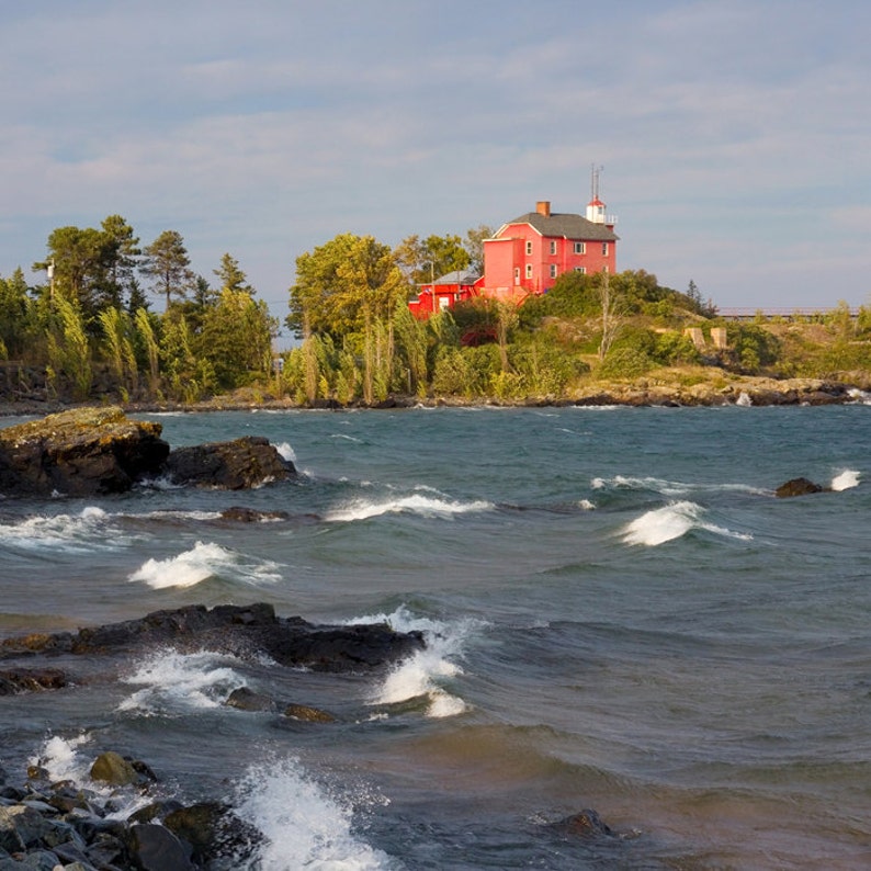 May include: A red lighthouse building on a small island with rocky shores and choppy water. The lighthouse is surrounded by trees and bushes.