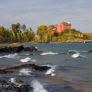 May include: A red wooden lighthouse sits on a rocky outcropping overlooking a choppy lake. The lighthouse has a white trim and a white roof. The lake is a deep blue color and the waves are white.