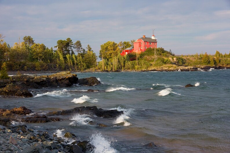 May include: A red wooden lighthouse sits on a rocky outcropping overlooking a choppy lake. The lighthouse has a white trim and a white roof. The lake is a deep blue color and the waves are white.