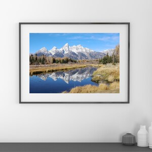 May include: A framed photograph of a mountain range reflected in a still lake. The mountains are snow-capped and the lake is a deep blue. The foreground is a grassy area with trees and bushes.