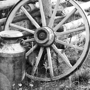 Puede incluir: Una foto en blanco y negro de una rueda de carro de madera vintage apoyada en una valla de madera. Una lata de leche de metal se encuentra delante de la rueda.