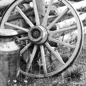 Puede incluir: Una foto en blanco y negro de una rueda de carro de madera vintage apoyada en una valla de madera. Una lata de leche de metal se encuentra delante de la rueda.