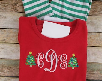 May include: A red long-sleeve shirt featuring an embroidered white monogram with the letters CDG flanked by two green Christmas trees with colorful ornaments and yellow stars, paired with green and white horizontally striped pajama pants.