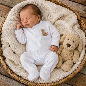 May include: A white long-sleeved footed sleeper with light blue trim, featuring an embroidered brown dog and the name Parker in light blue script. The sleeper is inside a woven basket with a cream-colored blanket and a plush tan dog toy.