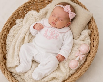 May include: A white long-sleeved footed sleeper with a pink Peter Pan collar and pink embroidered monogram on the chest. The sleeper is paired with a matching pink bow headband and a white and pink polka-dot rattle with a small pink bow.