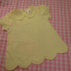 May include: A pale yellow short-sleeved shirt featuring a rounded Peter Pan collar, gathered puff sleeves, and a scalloped hemline. The garment includes a small metal snap closure at the back of the neckline and a white interior brand label.