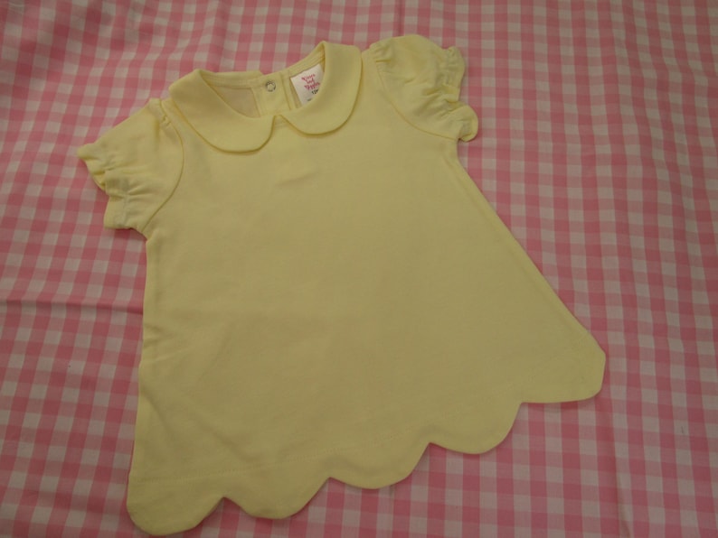 May include: A pale yellow short-sleeved shirt featuring a rounded Peter Pan collar, gathered puff sleeves, and a scalloped hemline. The garment includes a small metal snap closure at the back of the neckline and a white interior brand label.