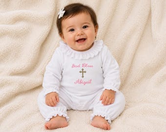 May include: A white long-sleeved infant romper with ruffled collar and cuffs. The chest features embroidered pink text reading God Bless and Abigail, separated by a gold-colored cross. A small white bow is attached to the hair of the infant.