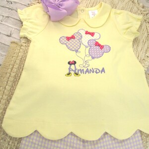 May include: A pale yellow short-sleeved shirt with a scalloped hem, featuring an embroidered design of three mouse-shaped balloons in purple and white gingham, each with a red bow, and the name Amanda embroidered in purple with mouse shoes.