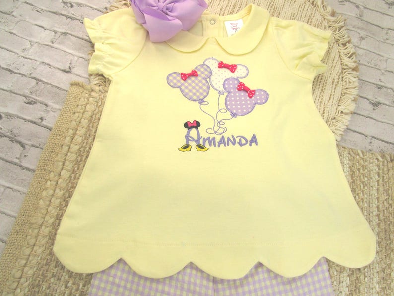 May include: A pale yellow short-sleeved shirt with a scalloped hem, featuring an embroidered design of three mouse-shaped balloons in purple and white gingham, each with a red bow, and the name Amanda embroidered in purple with mouse shoes.