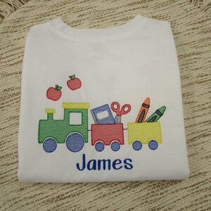May include: White long-sleeve shirt with a green, red, and yellow train carrying school supplies, including a book, scissors, and crayons. Two red apples are above the train. The name "James" is embroidered below the train.