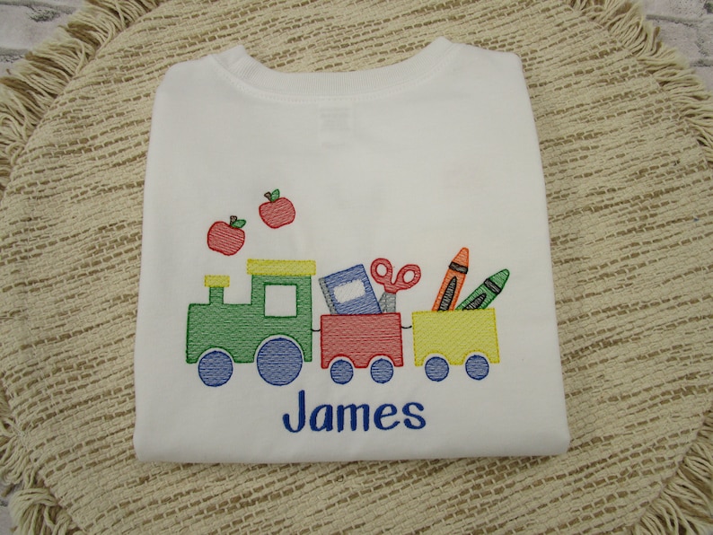 May include: White long-sleeve shirt with a green, red, and yellow train carrying school supplies, including a book, scissors, and crayons. Two red apples are above the train. The name "James" is embroidered below the train.