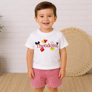 May include: A white short-sleeved t-shirt featuring the name Theodore in red text, surrounded by colorful cartoon icons including Mickey Mouse ears, gloves, shoes, and balloons, paired with red and white gingham patterned shorts.