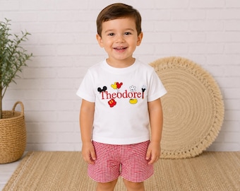 May include: A white short-sleeved t-shirt featuring the name Theodore in red text, surrounded by colorful cartoon icons including Mickey Mouse ears, gloves, shoes, and balloons, paired with red and white gingham patterned shorts.