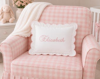 May include: A rectangular white throw pillow with a scalloped edge and the name Elizabeth embroidered in pink script, resting on a pink and white gingham upholstered armchair draped with a white knit blanket.