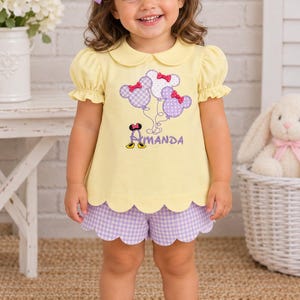 May include: A pale yellow short-sleeved shirt with a Peter Pan collar and scalloped hem, paired with light purple gingham shorts. The shirt features an embroidered design of three mouse-ear balloons and the name Amanda in purple lettering.