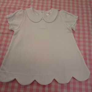 May include: A solid white short-sleeved shirt featuring a rounded Peter Pan collar, gathered sleeve cuffs, and a scalloped hemline. The garment includes a small metal snap closure at the back of the neckline and a visible interior brand label.